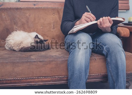 A young man is sitting on a sofa with a cat and is writing in a notebook
