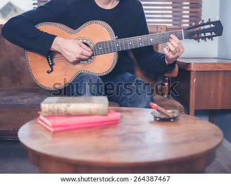 A young man is sitting on a sofa at home in his living room and is playing guitar, there is a coffee table with books on it in the foreground