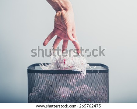A hand is putting a bunch of shredded paper in a waste paper basket