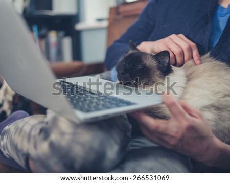 A young woman is using her laptop at home with a cat sitting on her lap