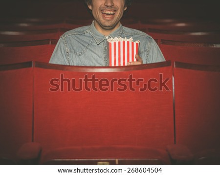 A happy young man with a bucket of popcorn is laughing and watching a movie at the cinema