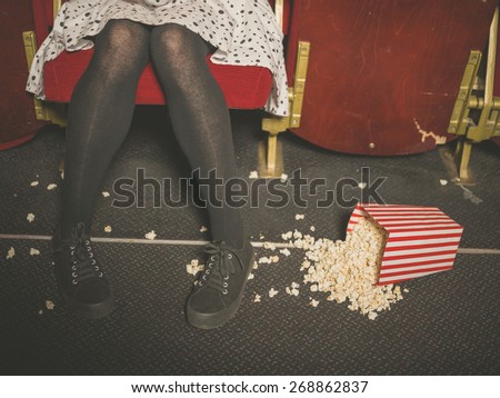 A young woman is sitting in a movie theater with a bucket of popcorn spilled on the floor in front of her