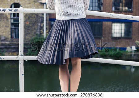 A young woman wearing a skirt is standing on a bridge by a canal and is relaxing whilst looking at an old building