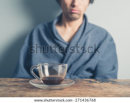 A tired young man wearing a bathrobe is sitting at a table and is trying to wake up by having a cup of coffee