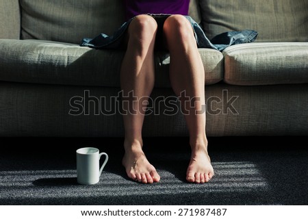 A woman is sitting on a sofa with a mug on the floor next to her feet
