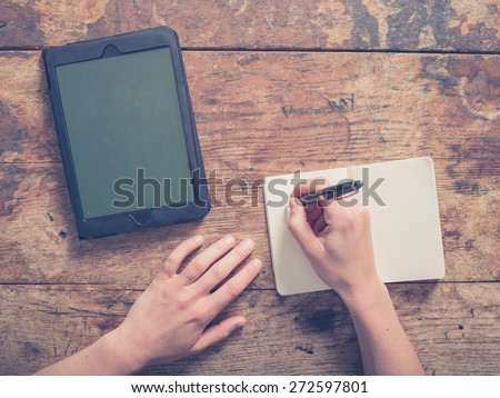 Close up on a woman writing in a small notepad with a tablet computer next to her hands on a wooden table