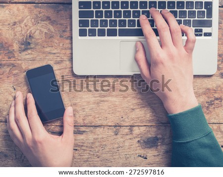 Close up on the hands of a young man as he is using a laptop and a smart phone at a wooden table