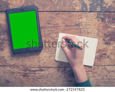 A male hand is writing in a notepad there is a tablet computer on the table as well with a green screen