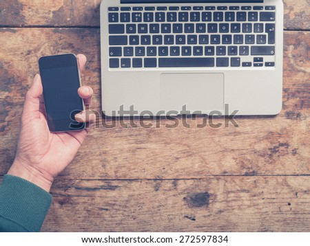 Close up on the hands of a young man as he is using a laptop and a smart phone at a wooden table