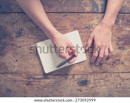 Close up on the hands of a young woman as she is writing in a small notepad at a wooden table
