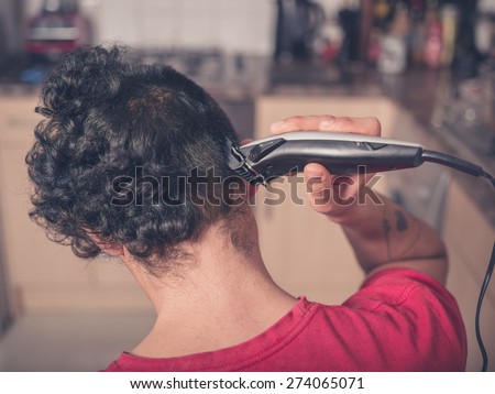 A young man is using hair clippers to give himself a haircut in his kitchen