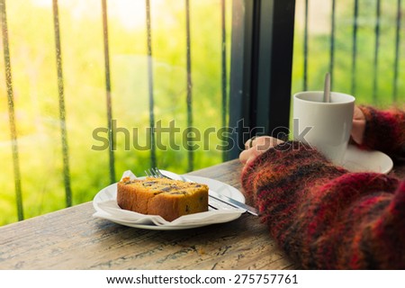The hands of a young woman resting on a table by the window as she is having tea and cake