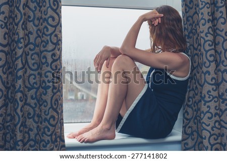 A young woman is sitting on a window sill and is looking out on a rainy day