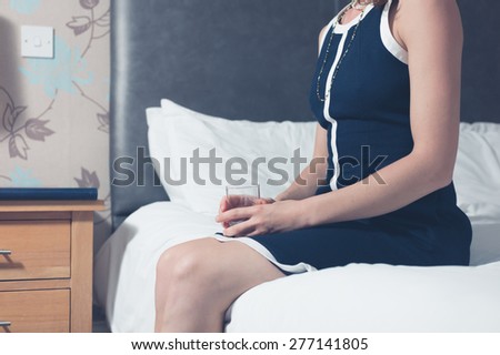 An elegant young woman is sitting on a bed in a hotel room with a glass in her hand