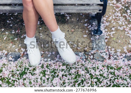 The legs and feet of a young woman relaxing on a park bench with cherry blossom on the ground