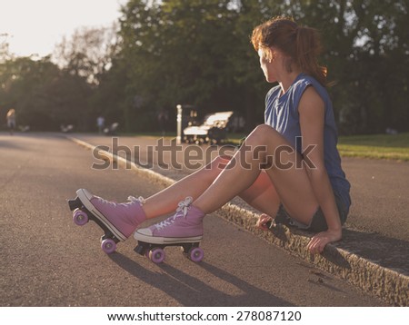 A young woman is sitting on the ground in a park at sunset and she is wearing roller skates