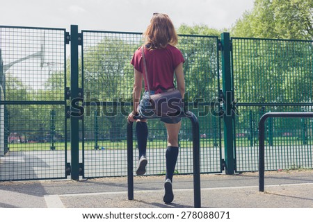 A young woman is sitting on a rail in a park outside a basketball court on a summer day