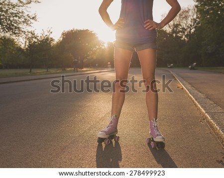 An attractive young woman wearing roller skates is standing in a park admiring the sunset
