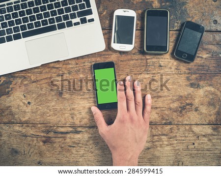 Overhead shot of a male hand using a smart phone with a green screen on a wooden table. There is a laptop and other smart phones on the table too.