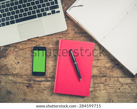 Overhead shot of a smart phone with a green screen surrounded by a book, a laptop, a notepad and a pen