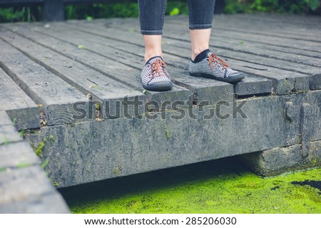 The feet of a young woman standing on a wooden deck by a dirty pond with algae