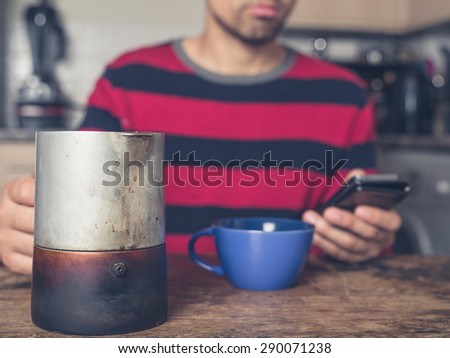 A young man is sitting in a kitchen and is drinking coffee while using a smart phone
