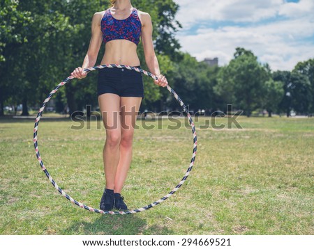 A fit young woman is standing on the grass in a park on a sunny summer day and is holding a hula hoop