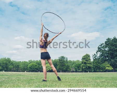 A fit and athletic young woman is standing on the grass in a park with a hula hoop raised above her head