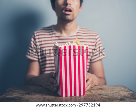 A young man with a stunned look on his face is sitting at a table with a box of popcorn