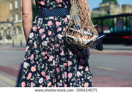A young woman wearing a dress is standing in the street with a net bag containing a pair of spare party shoes