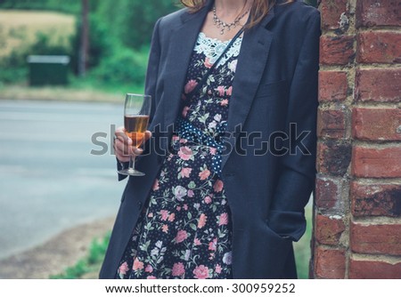 A young woman wearing a man\'s jacket over her dress is standing with a drink by a wall in the countryside