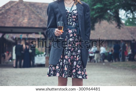 A young woman wearing a man\'s jacket over her dress is standing with a drink at a party in the countryside