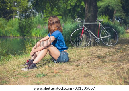 A young woman is relaxing by the water in a park on a summer day  with her bicycle resting against a tree in the background