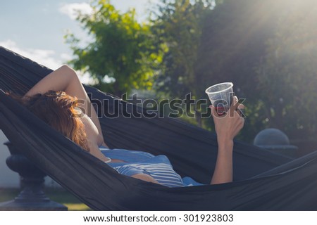 A young woman with a drink in her hand is relaxing in a hammock outside in a garden on a sunny summer day