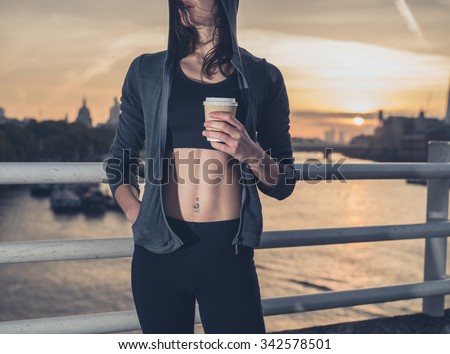 An athletic young woman with toned abs  is standing on a bridge in London at sunrise with a paper cup in her hand