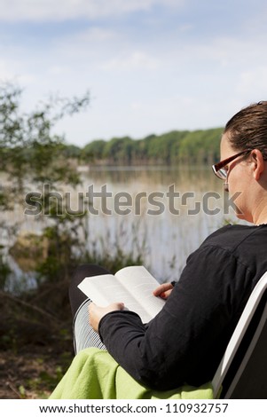 Female sitting in a chair reading a book with a lake in front of her. A boat is sailing on the lake. Focus is on the boat.