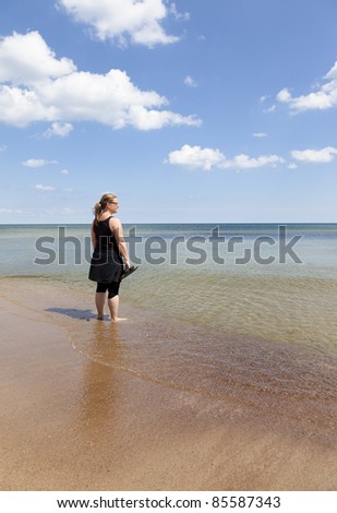 Woman standing with feet in the water watching towards the horizon. Sky is blue with tiny clouds