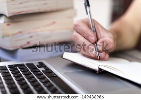Young traveler writing down journey stories in a notebook with a laptop and books in the background