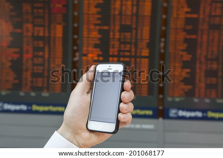 Checking smartphone screen at flight information board timetable in airport