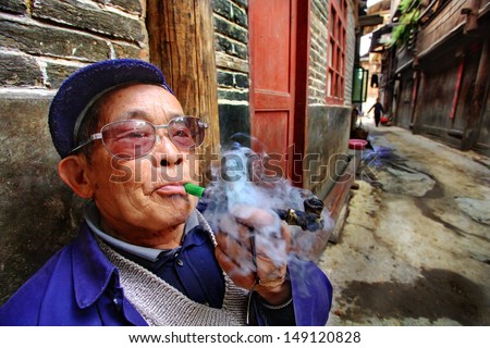 GUIZHOU, CHINA - APRIL 9:  An elderly Chinese farmer wearing glasses, smoking a pipe outside his home April 9, 2010. Zhaoxing Dong villages, Liping County. Chinese peasant produces tobacco smoke.