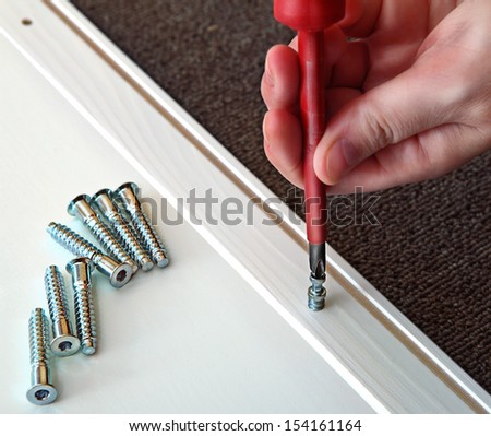 Hand holds cruciate screwdriver with a red handle, screwing wood dowel is a wooden panel, covered with a white stain. On the panel are furniture screws.