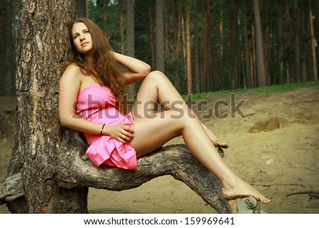 Teen girl 16 years old, Caucasian appearance, in pink dress, rest on nature, in  pine forest. Slender girl with long brown hair and bare feet, relaxing in a forest clearing, sitting on a pine branch.