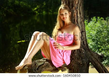 Girl caucasian appearance, 16 years old, in a pink dress, with long hair and bare feet, sitting on a tree trunk, near the dark waters of a forest lake.