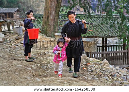 GUIZHOU PROVINCE, CHINA - APRIL 11:  Asian village, Chinese woman goes fetch water, she carries pails on yoke, and leads by hand of child, girl 3 years old, a child holding a toy, April 11, 2010.