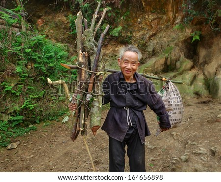 GUIZHOU PROVINCE, CHINA - APRIL 11: An elderly Chinese man goes on mountain road with load on yoke, carries nylon bag and an armful of branches for  hearth, Zengchong village,  - April 11, 2010.