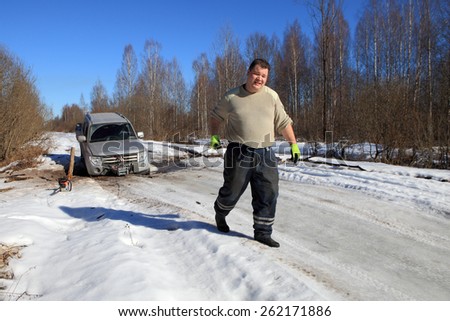 Nazia village, Leningrad Region, Russia - March 17, 2015: Car stuck on a forest road, front wheel fell into ravine in the ice, the man pulls the cable car winch.