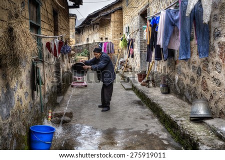 Yang Di village, Yangshuo, Guangxi, China - March 29, 2010: Old farmer pours water from a bucket on a narrow street in a peasant village, close to  rope hung to dry clothes. Guangxi Zhuang region.