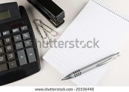 A desktop calculator, stapler and paper clips stands next to a blank writing pad with a stylish pen atop