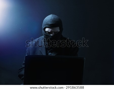 Angry computer hacker in suit stealing data from laptop in front of black background and blue light
