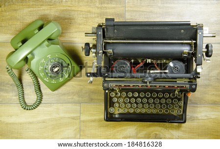 Old rotary phone next to a typewriter on a wood table.